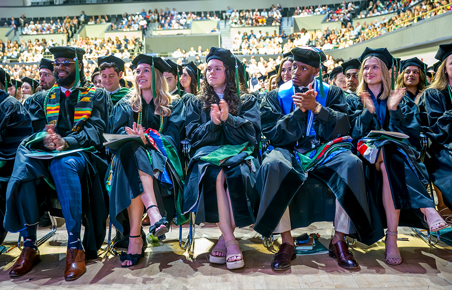 A group of medical students in full graduation regalia clap during a large ceremony.