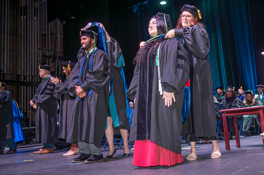 Two medical school graduates, one man and one woman, have their ceremonial hoods placed around their shoulders during a hooding ceremony.