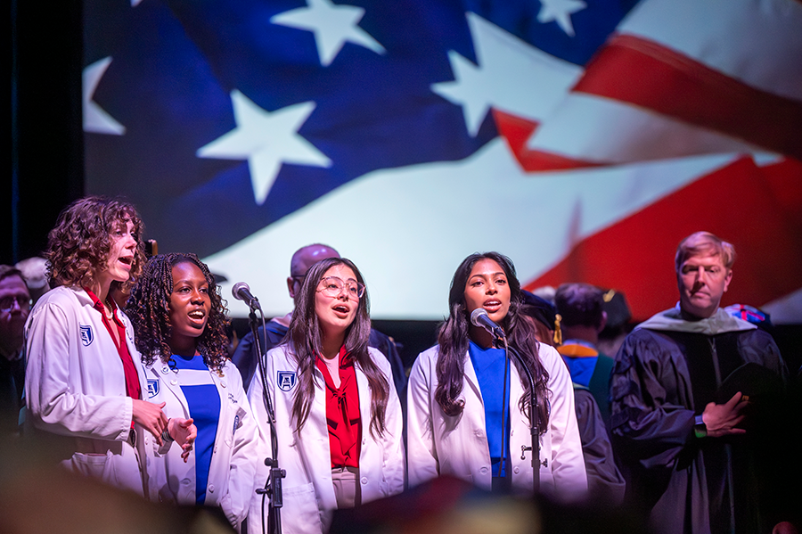 Four female medical students, all wearing lab coats over dress clothes, sing the U.S. national anthem.