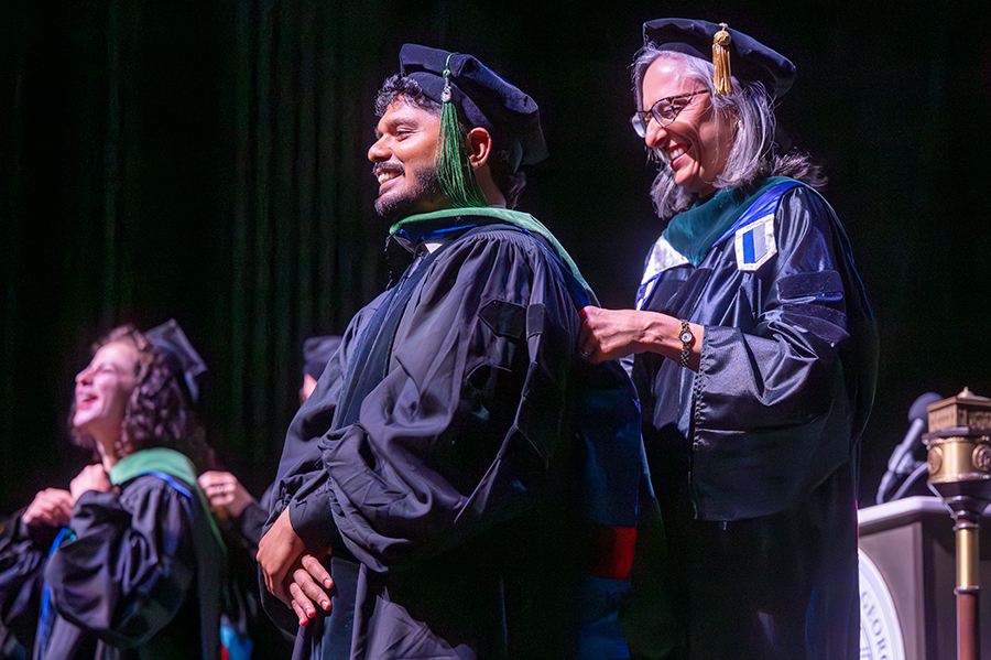 A male medical student in full graduation regalia smiles as a professor places a ceremonial hood around his shoulders during a graduation ceremony.