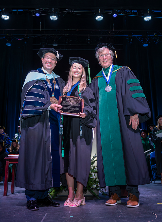 Two men, both wearing full college graduation regalia, present a woman, also wearing full regalia, with an award during a college hooding and commencement ceremony.