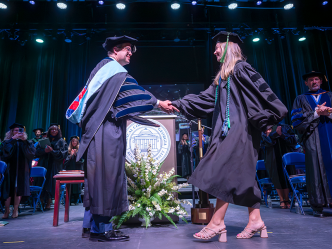 A woman wearing full graduation regalia walks across a graduation stage and shakes the hand of a college president, also dressed in full graduation regalia. The president is presenting the woman with a special award in front of a large crowd.