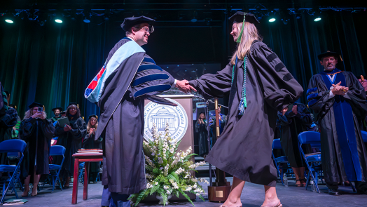 A woman wearing full graduation regalia walks across a graduation stage and shakes the hand of a college president, also dressed in full graduation regalia. The president is presenting the woman with a special award in front of a large crowd.