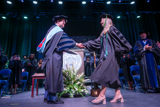 A woman wearing full graduation regalia walks across a graduation stage and shakes the hand of a college president, also dressed in full graduation regalia. The president is presenting the woman with a special award in front of a large crowd.