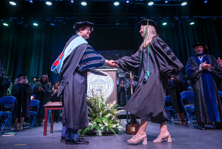A woman wearing full graduation regalia walks across a graduation stage and shakes the hand of a college president, also dressed in full graduation regalia. The president is presenting the woman with a special award in front of a large crowd.