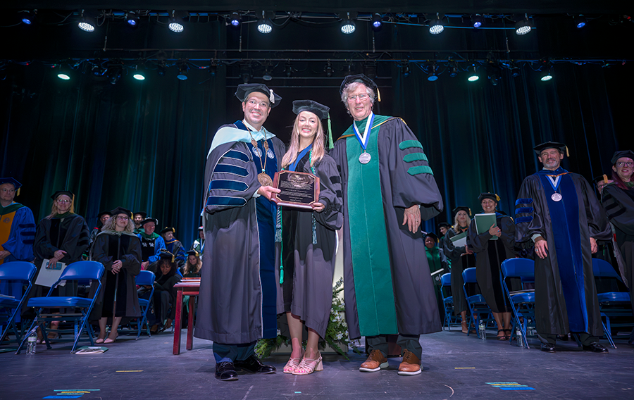 Two men, both wearing full college graduation regalia, present a woman, also wearing full regalia, with an award during a college hooding and commencement ceremony.