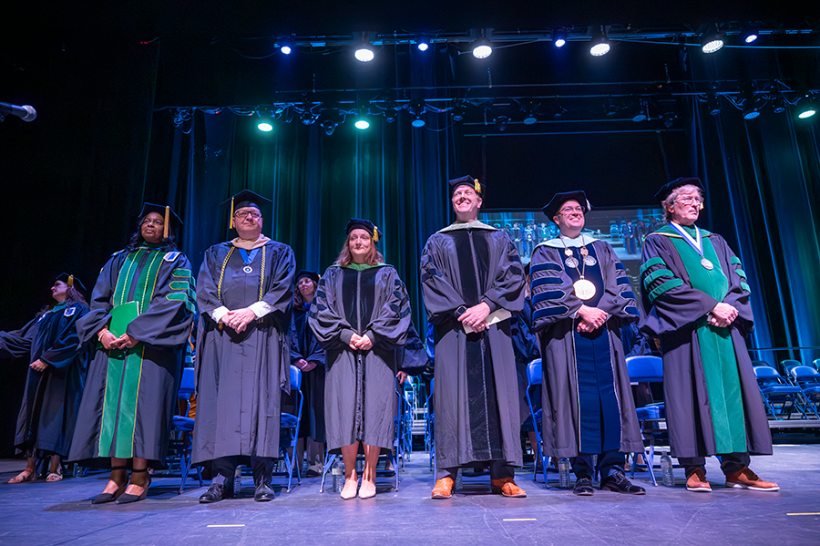 Six university professors and staff stand on a stage in full graduation regalia during a graduation ceremony in a large auditorium.