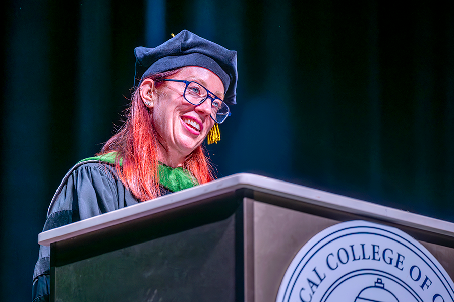 A woman in full medical school graduation regalia stands at a podium and delivers a keynote address to a large crowd.