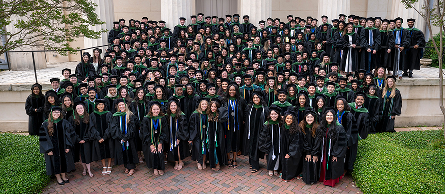 Over 200 medical school graduates in full graduation regalia stand in a large group on the steps of an old building.