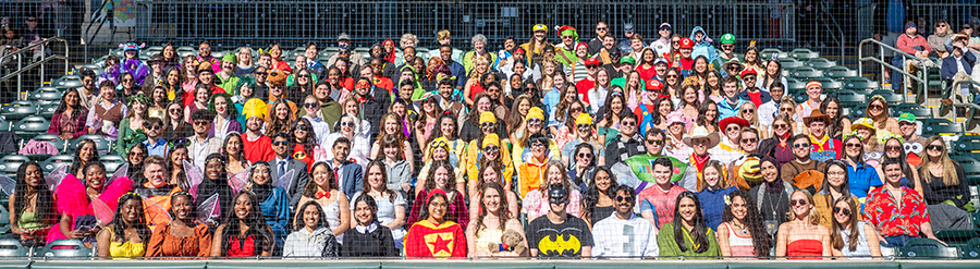 A large group of over 150 medical students sit in the stands of a baseball stadium. They are all dressed up in costumes of different heroes and villians.