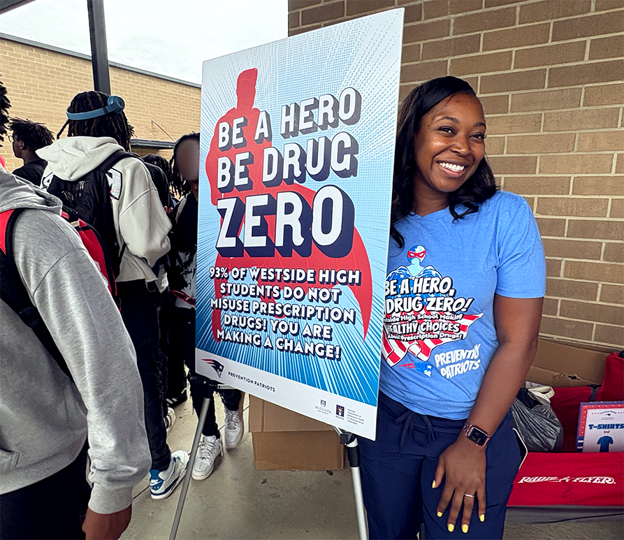 A woman wearing a shirt that has "Be a Hero, Be Drug Zero!" printed on it poses beside a sign that reads the same thing.