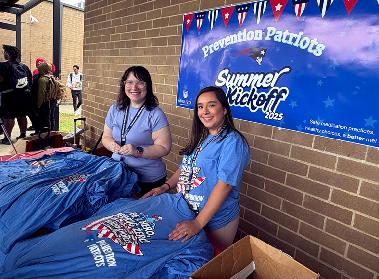 Two women smile while handing out t-shirts advocating for drug misuse prevention.