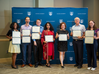 A group of seven college faculty members, including four women and three men, hold up special awards during an award ceremony in a large banquet hall.