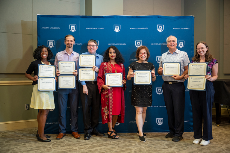 A group of seven college faculty members, including four women and three men, hold up special awards during an award ceremony in a large banquet hall.