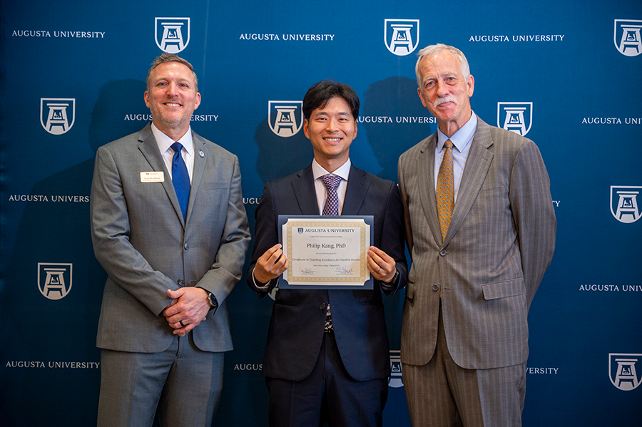 Three men stand in a large ballroom. The man in the middle is holding a certificate for completing a training program.