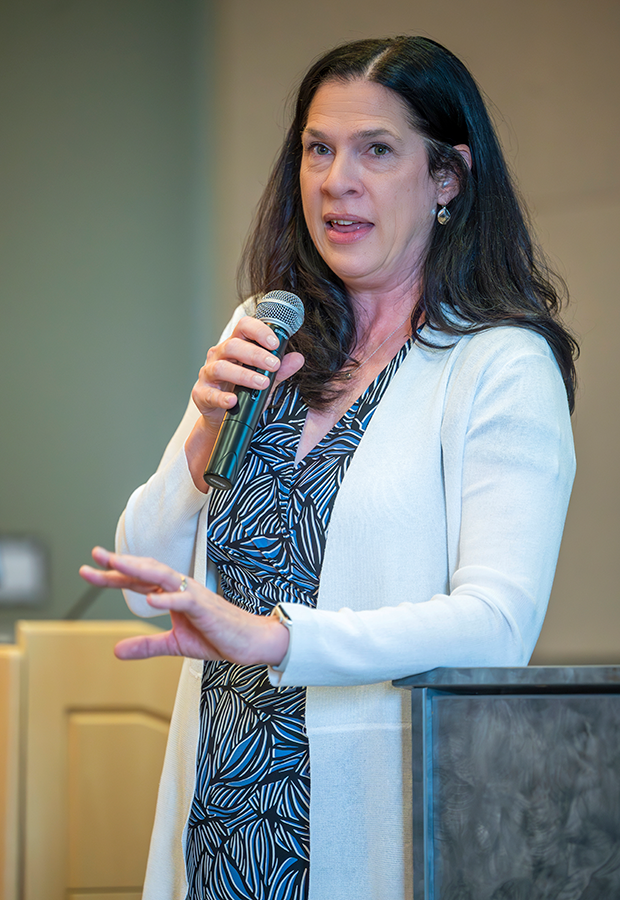 A woman holds a microphone and speaks to a large group of university faculty gathered for a celebration in a large ballroom.