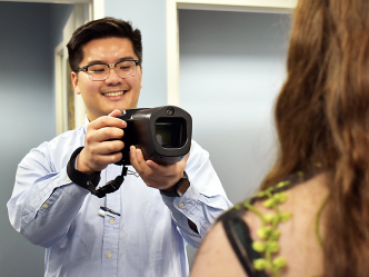 A male college student holds a camera and points it towards a woman to take a picture of her eyes for a medical screening.