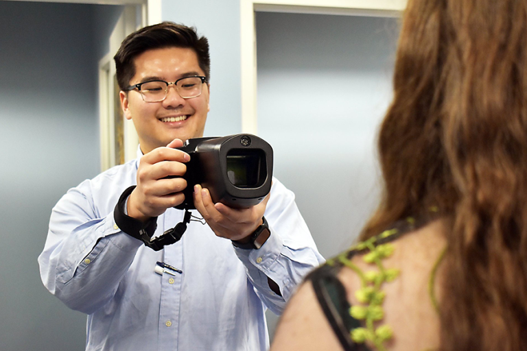 A male college student holds a camera and points it towards a woman to take a picture of her eyes for a medical screening.