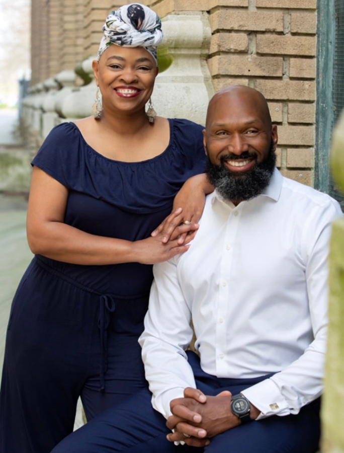 A standing woman props her arm on the shoulders and leans on a man who is sitting in front of a brick building.
