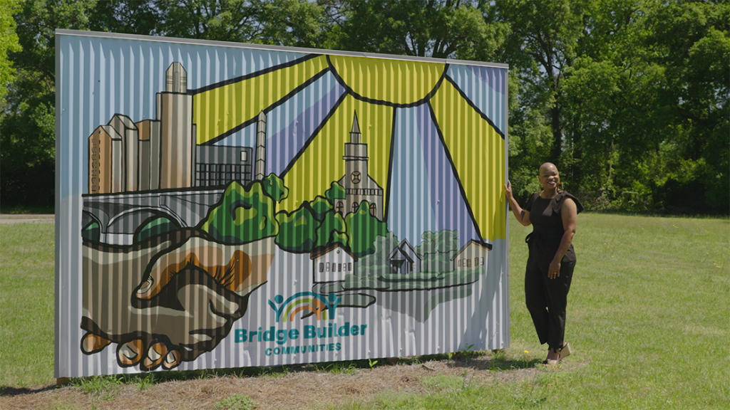 Woman stands in front of a mural painted on a metal structure, in front of lots of trees.