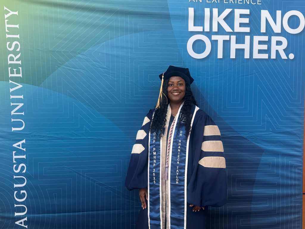 A woman in full doctoral regalia poses in front of a blue Augusta University step and repeat