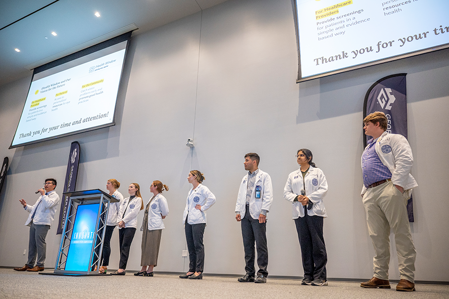 A group of 8 medical students stand at the front of a large lecture hall. One of the students is holding a microphone to talk about their research project.