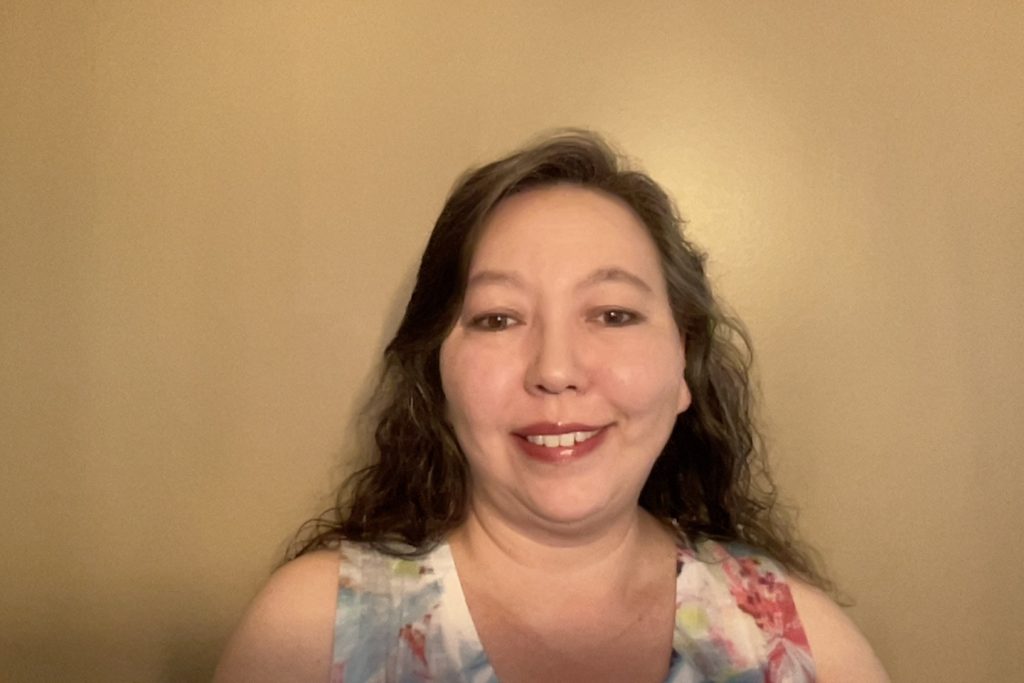 Woman in floral top looks at camera as she poses against a yellowish-tan wall