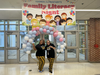 Two women, both elementary school teachers, stand in the entryway to a school under a balloon arch and a banner that reads "Family Literacy Night."