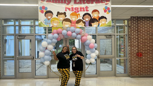 Two women, both elementary school teachers, stand in the entryway to a school under a balloon arch and a banner that reads "Family Literacy Night."