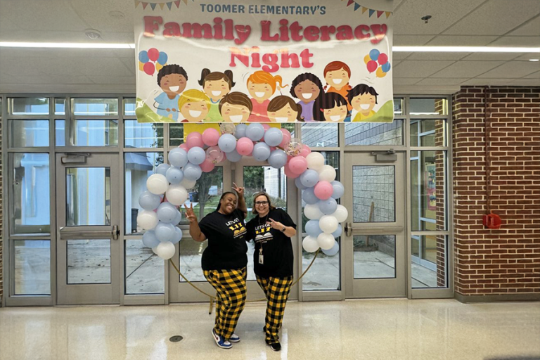 Two women, both elementary school teachers, stand in the entryway to a school under a balloon arch and a banner that reads "Family Literacy Night."