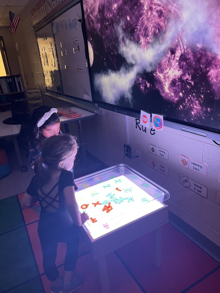 Two elementary school students work on an activity at a table illuminated by glowing alphabet letters.
