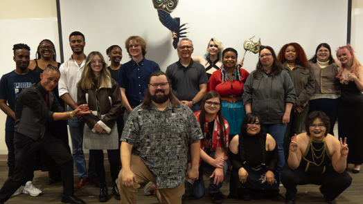A large group of college students with a male professor stand at the front of a classroom during a question and answer session.