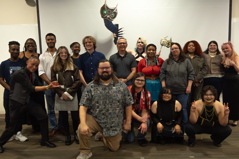 A large group of college students with a male professor stand at the front of a classroom during a question and answer session.
