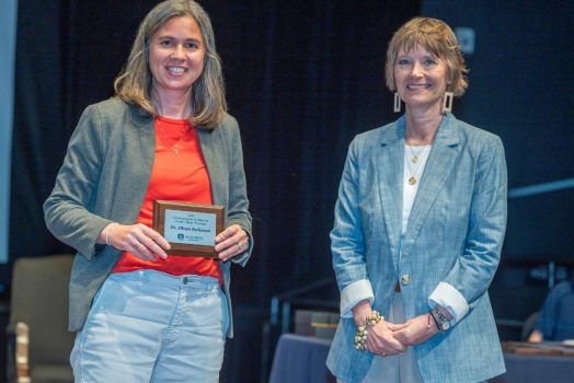 Two woman stand smiling at the camera. The woman on the left is holding a small plaque.
