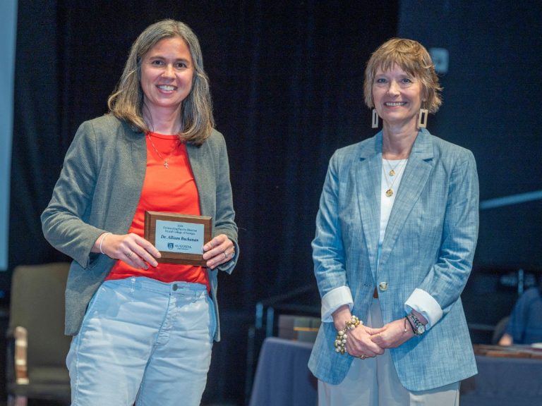 Two woman stand smiling at the camera. The woman on the left is holding a small plaque.