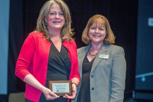Two women stand next to each other and smile at the camera. The woman on the left is holding a small plaque.