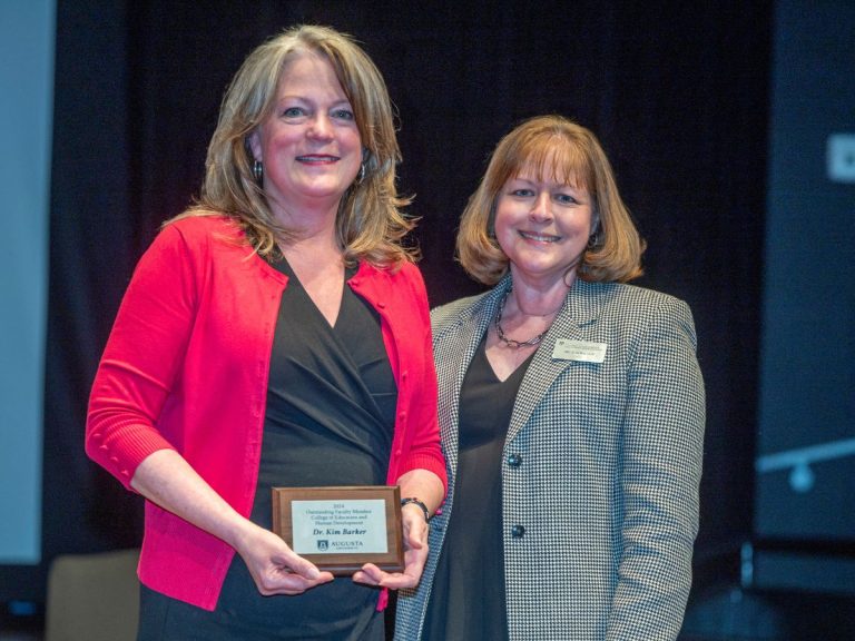 Two women stand next to each other and smile at the camera. The woman on the left is holding a small plaque.