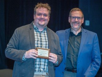 Two men stand next to each other smiling for the camera while one of the men holds a small plaque
