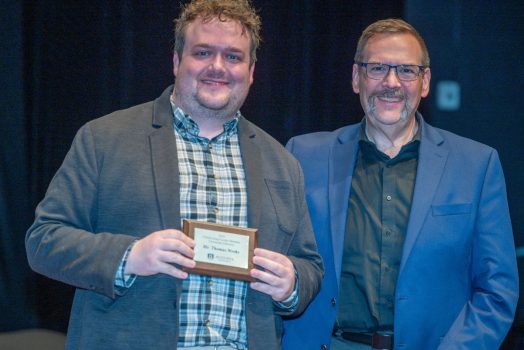 Two men stand next to each other smiling for the camera while one of the men holds a small plaque