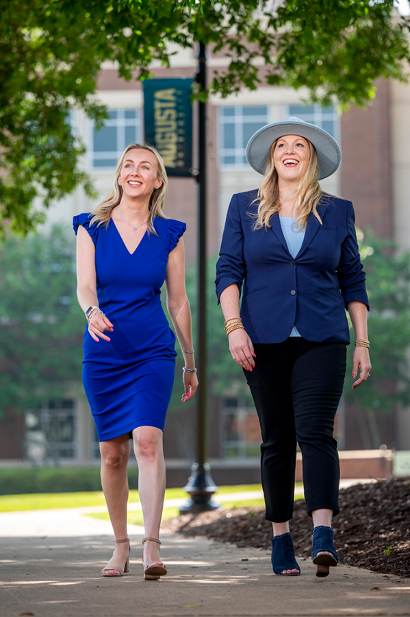 Two ladies walk on sidewalk next to each other. One is wearing a hat.