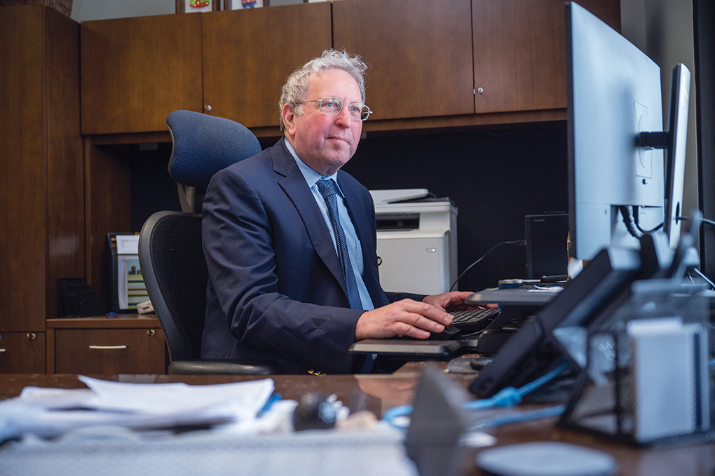 A doctor wearing a suit and tie sits at a desk and looks at a computer monitor.