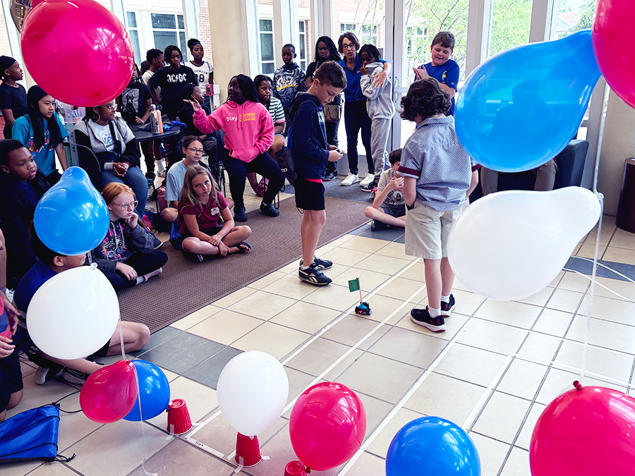 A crowd of young kids watch two young boys drive a remote-controlled miniature car through an obstacle race track.