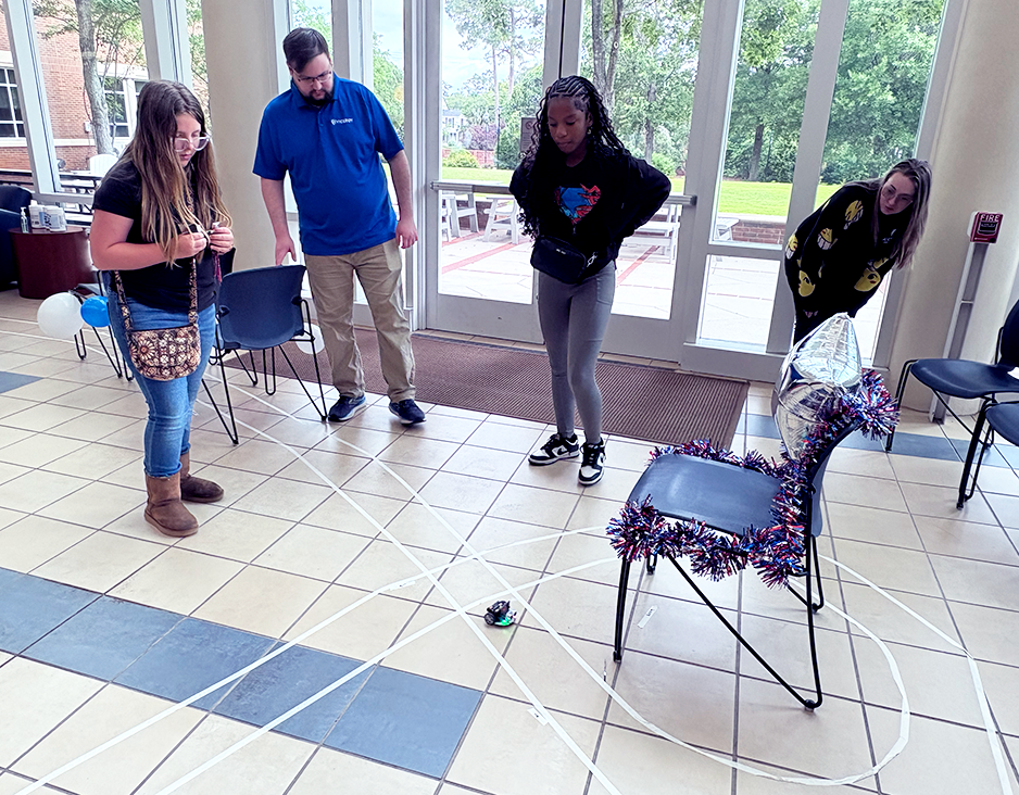 A young girl operates a remote-controlled miniature car through a race course while her teammate and adult teachers observe.