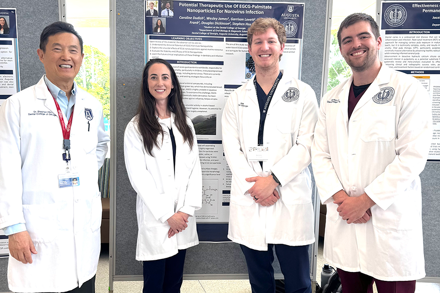 Three men and one woman, all wearing scientific lab coats, stand in front of a scientific poster during a special presentation.