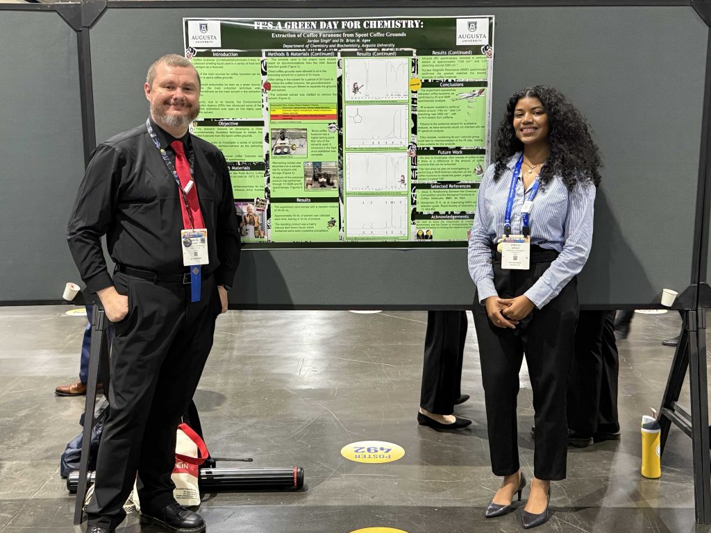 A man in a dark shirt and pants, stands with a woman in a blue shirt and dark pants in a conference center. The woman's poster presentation is between the two.