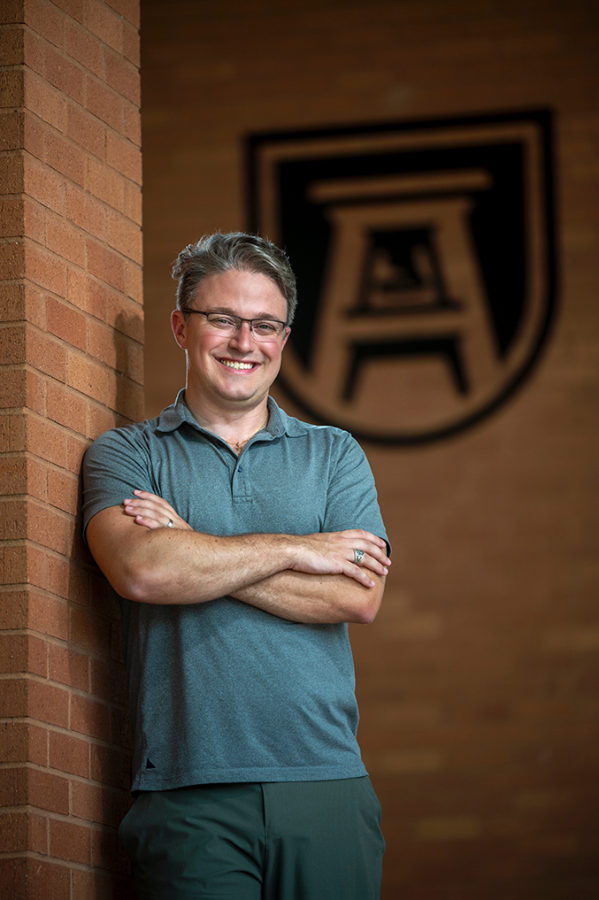 A man wearing glasses leans against a brick wall with his arms crossed.