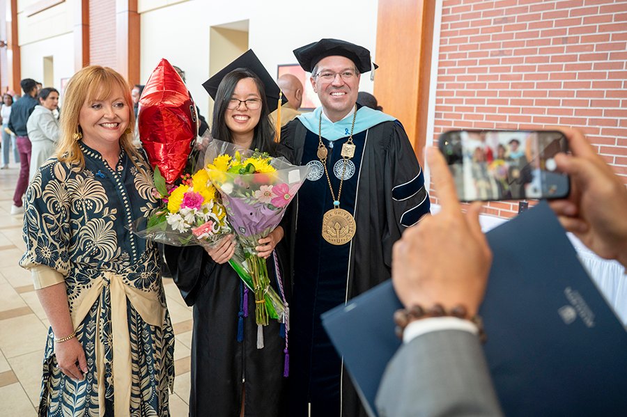 A college graduate wearing full graduation regalia and holding a bouquet of flowers poses for a photo with the university president, also in full graduation regalia, and the president's wife. The graduate's father holds up a cellphone to take their photo.