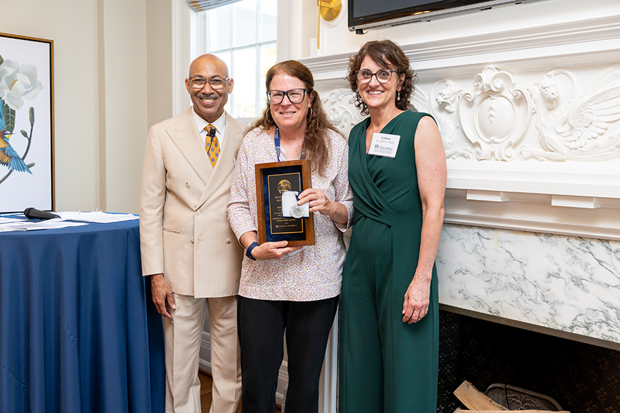A man and a woman stand beside another woman, who is smiling and holding a special plaque celebrating her being named a distinguished alumna.
