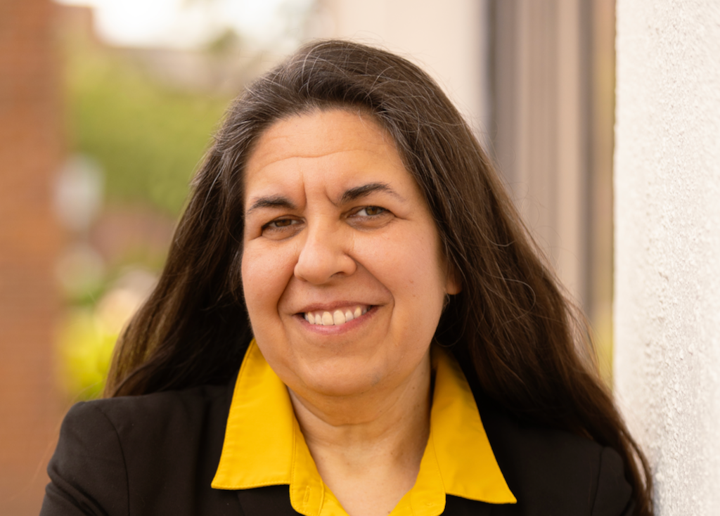 A woman wearing a collared yellow shirt and black blazer stands outside smiling at the camera.