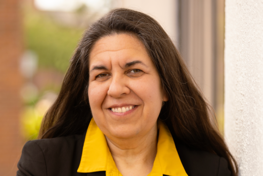 A woman wearing a collared yellow shirt and black blazer stands outside smiling at the camera.